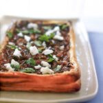 A white rectangular platter with a puff pastry tart filled with caramelized onions, feta cheese, and oregano.