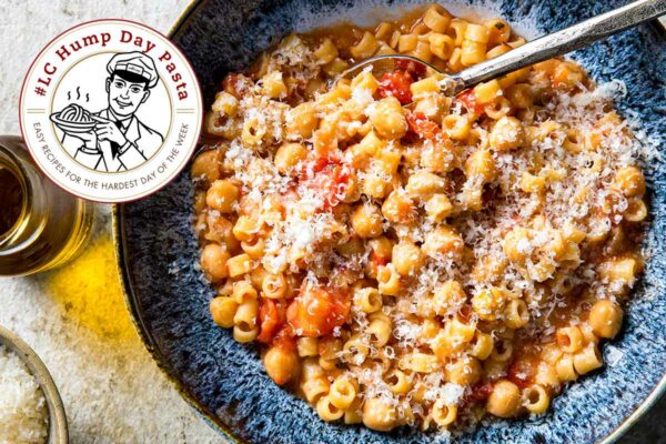 A blue bowl filled with chickpeas, diced tomatoes, ditalini pasta, and parmesan, flanked with a crueset of olive oil and a bowl of Parmesan.