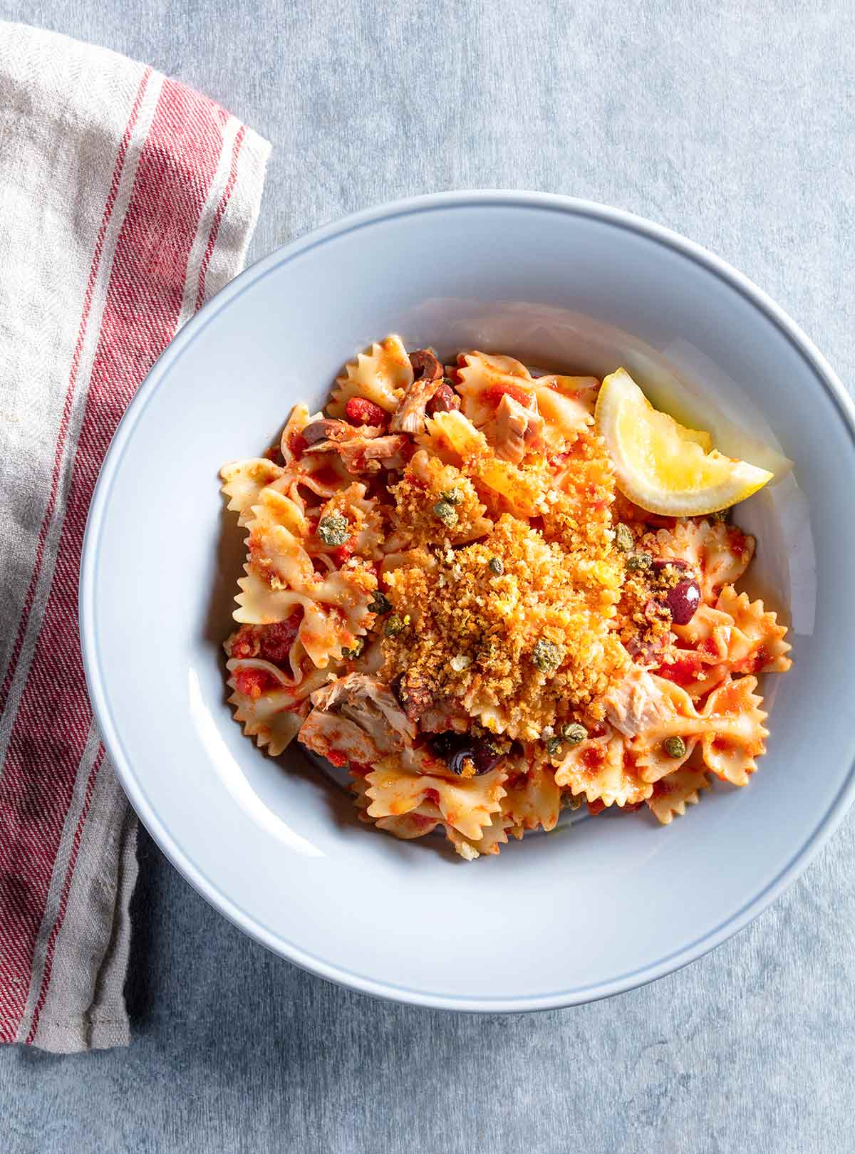 A pale blue bowl filled with farfalle, tuna, capers, tomatoes, and topped with bread crumbs and a slice of lemon, sitting on a red and white dish cloth.