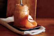 A jar of roasted applesauce with a spoon resting inside on a saucer with a strip of apple peel.