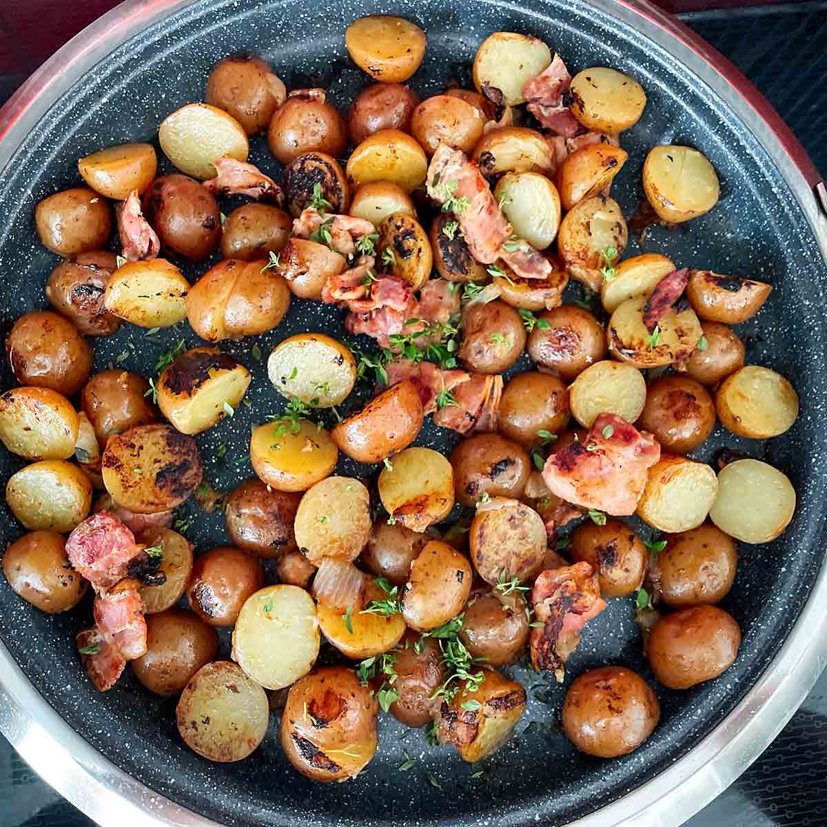 An enamelware bowl filled with braised, halved red potatoes, covered with bacon, onions, and parsley.