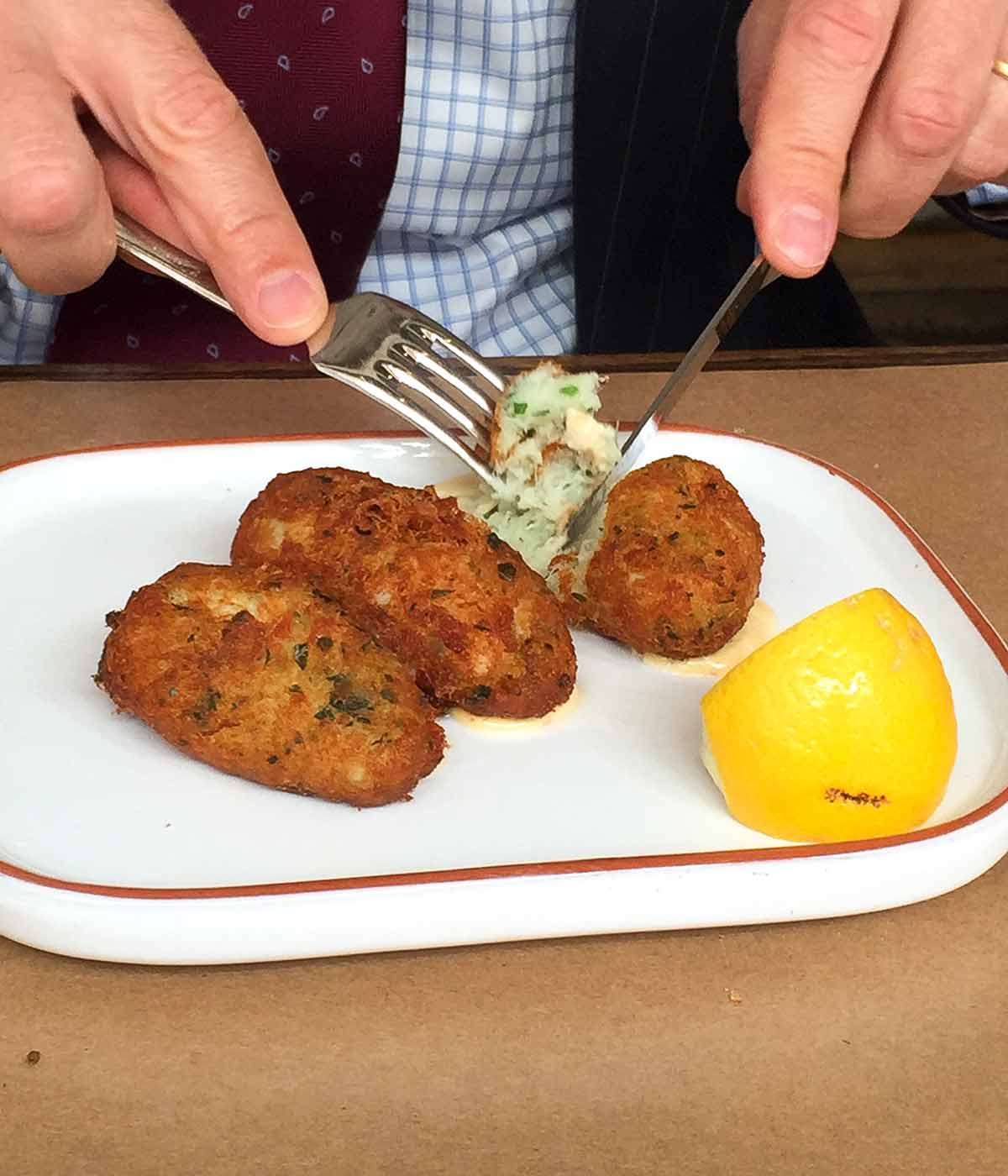 A close up of an oval plate with 3 salt cod fritters and a lemon wedge, one is being cut into with a knife and fork.
