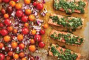 A metal sheet pan with 4 salmon fillets covered with gremolata, beside a pile of cherry tomatoes and diced red onions.