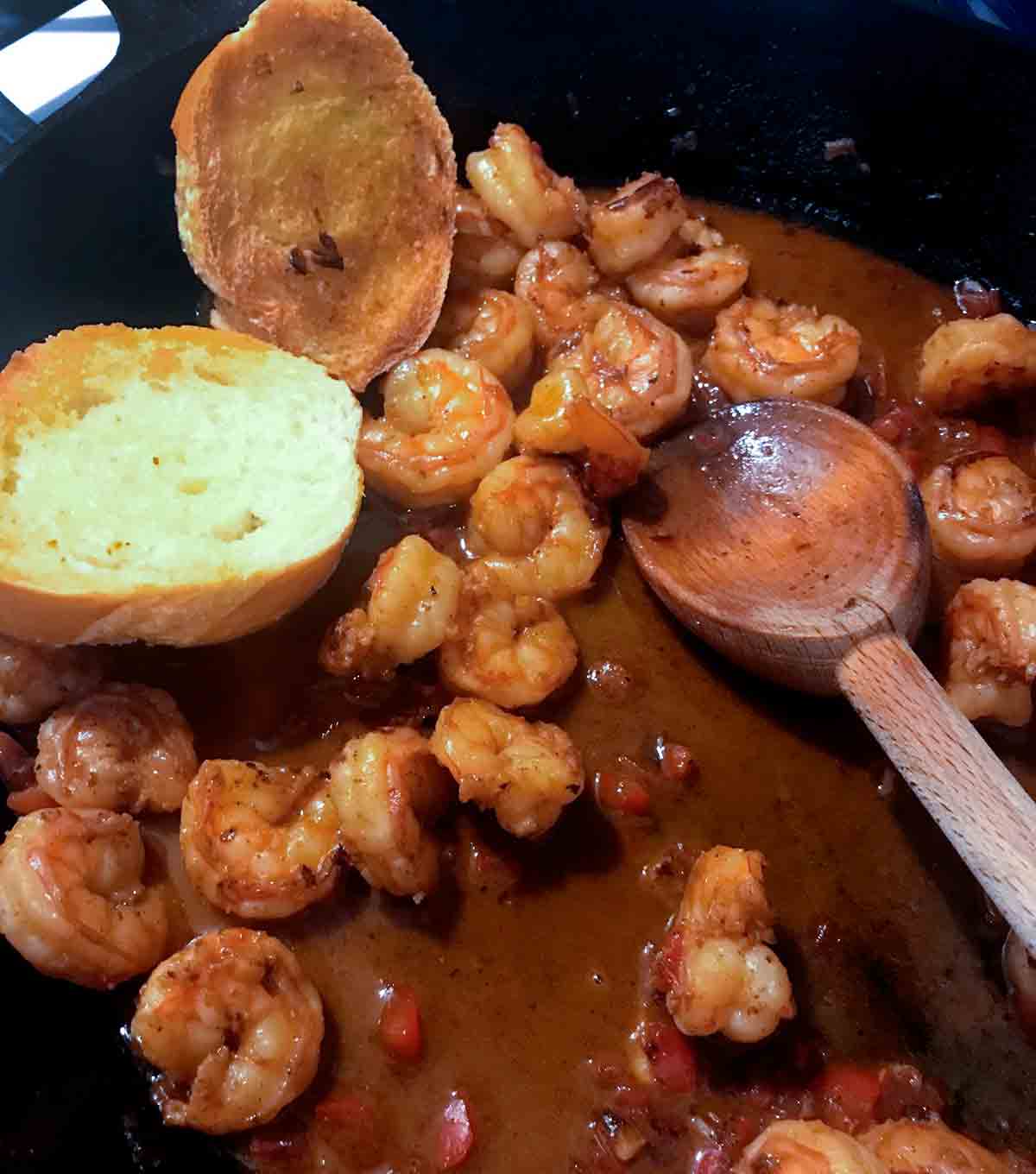 A cast-iron skillet filled with shrimp in a dark red sauce, with a wooden spoon and 2 pieces of toasted bread.