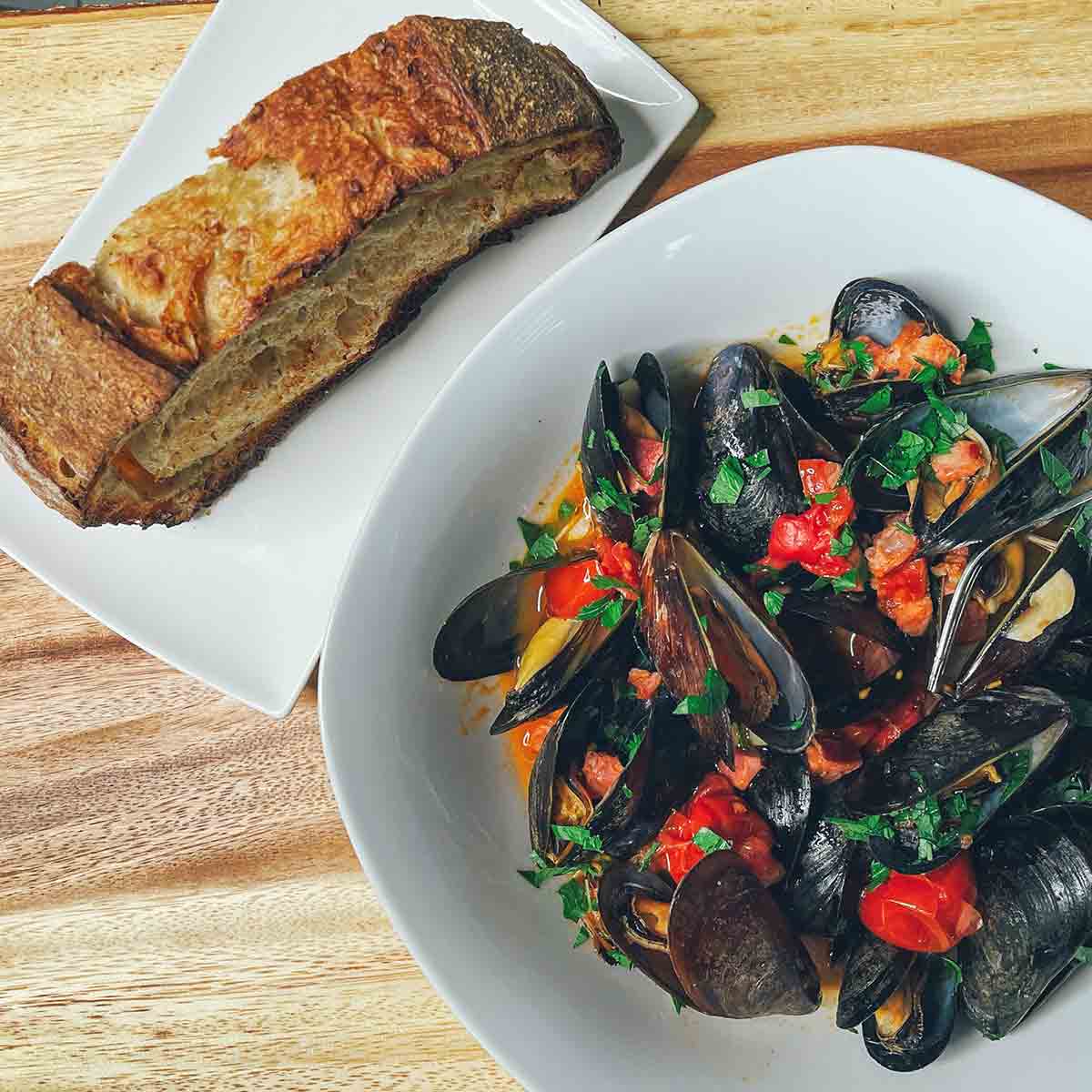 A white dish with a slice of crusty bread beside a white bowl filled with mussels, chorizo, peppers, and parsley.