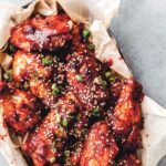 A basket of chicken wings with wax paper, covered with a sticky red sauce, sesame seeds, and green onion slices.
