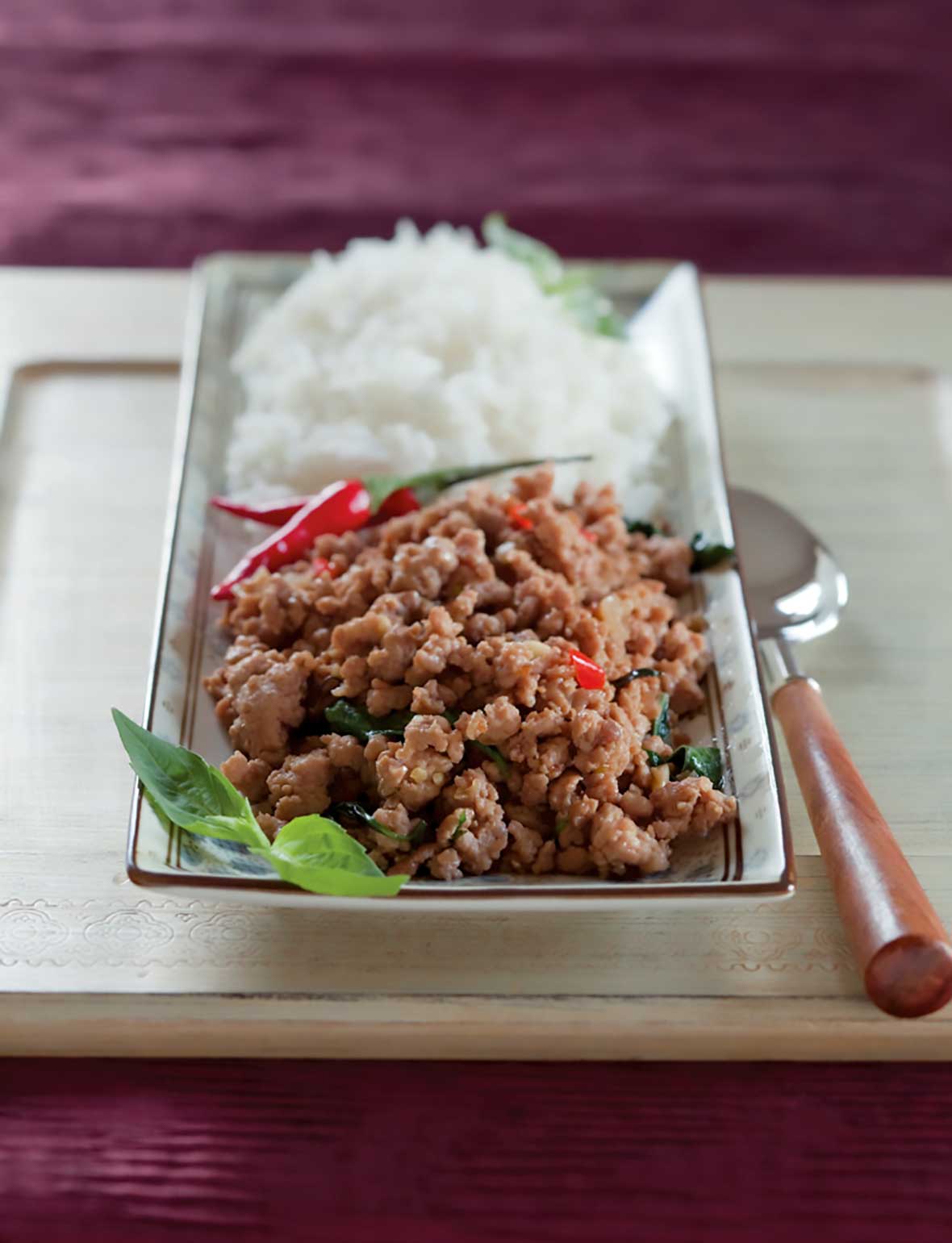 A wooden cutting board with a rectangular plate and spoon on top, the plate has a mound of rice on the back half. On the front half, there's a pile of ground pork, basil, and peppers.