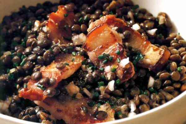 A creamy pottery bowl filled with a warm lentil salad, made with green and brown lentils, bacon, shallots and chives.