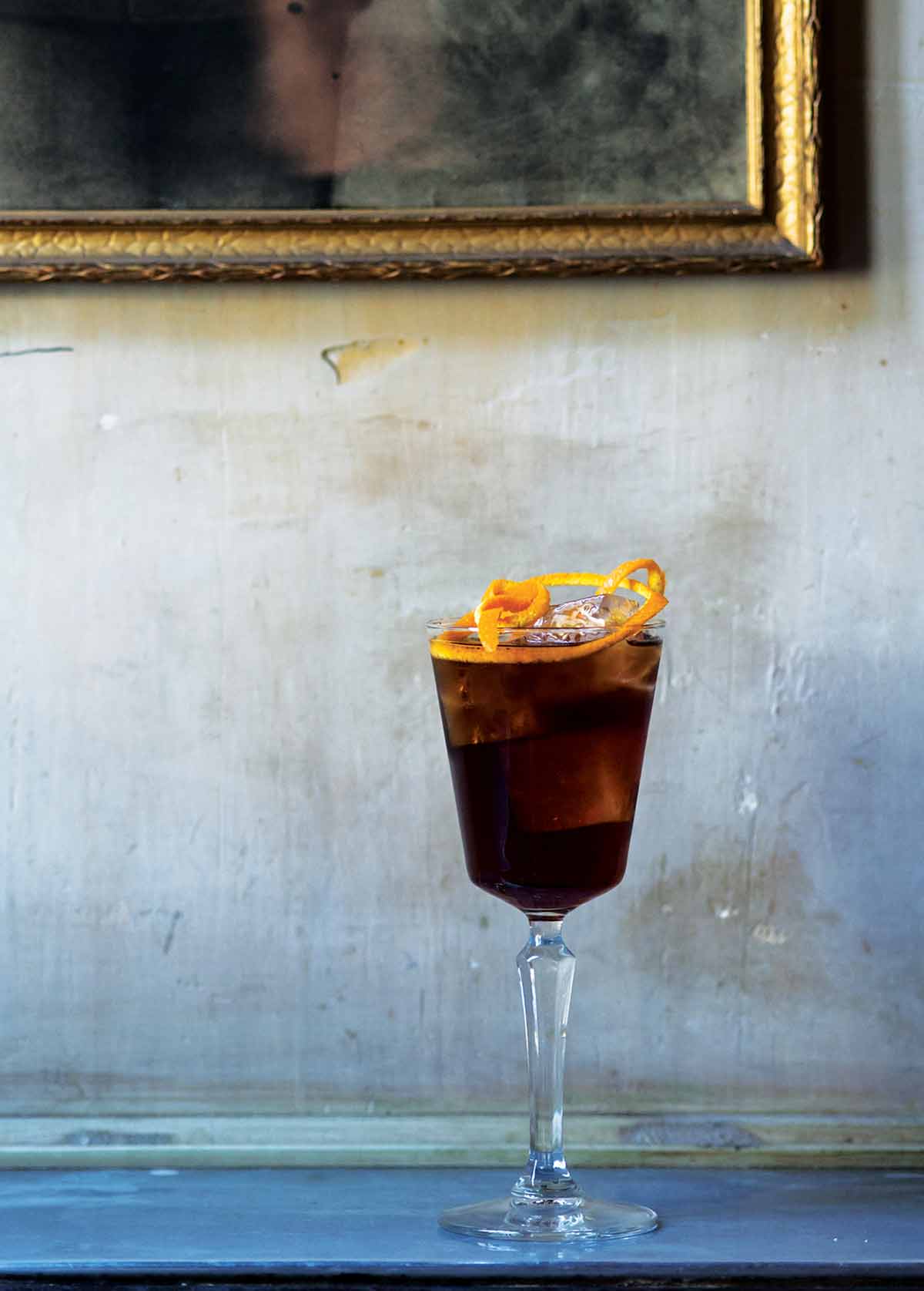 A glass gobelet filled with an Adonis cocktail and an orange twist garnish, in front of a painted background.