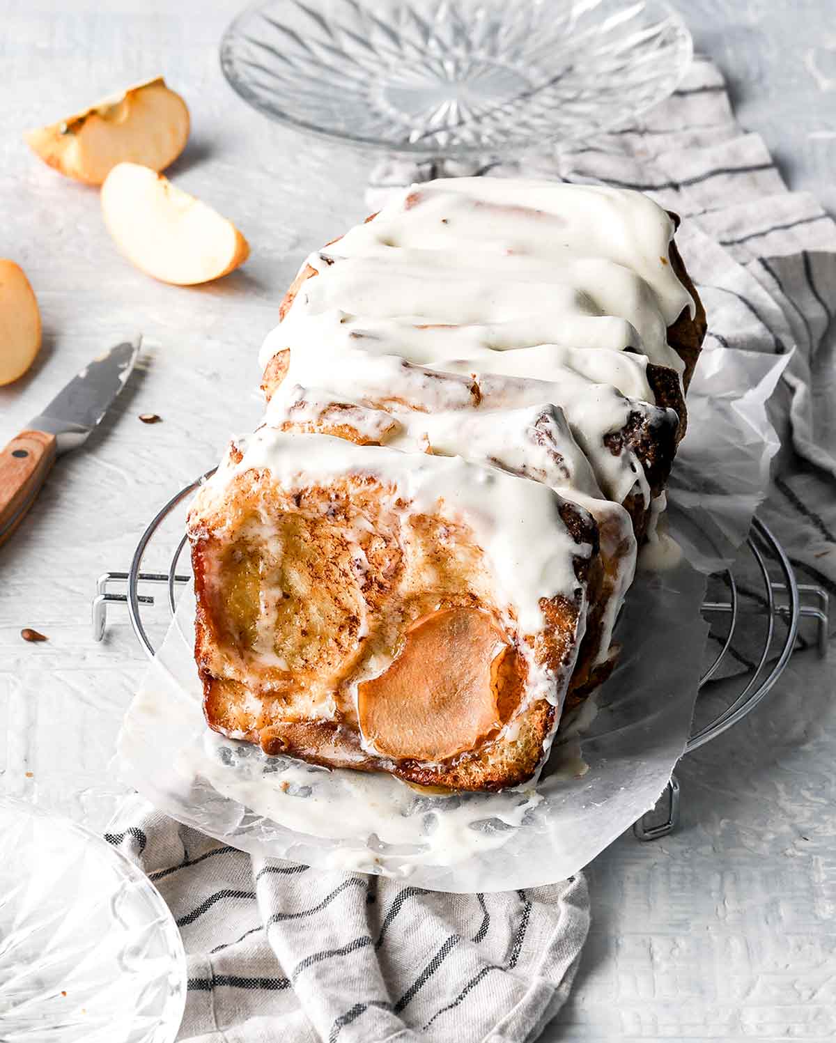 An apple cinnamon pull-apart bread on a piece of parchment paper on top of a wire cooling rack, with apple slices, a glass plate, and a knife in the background.