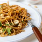 A white plate with sesame noodles, garnished with cilantro, peanuts, and sesame seeds, with a fork and a bowl of green onions and soy.
