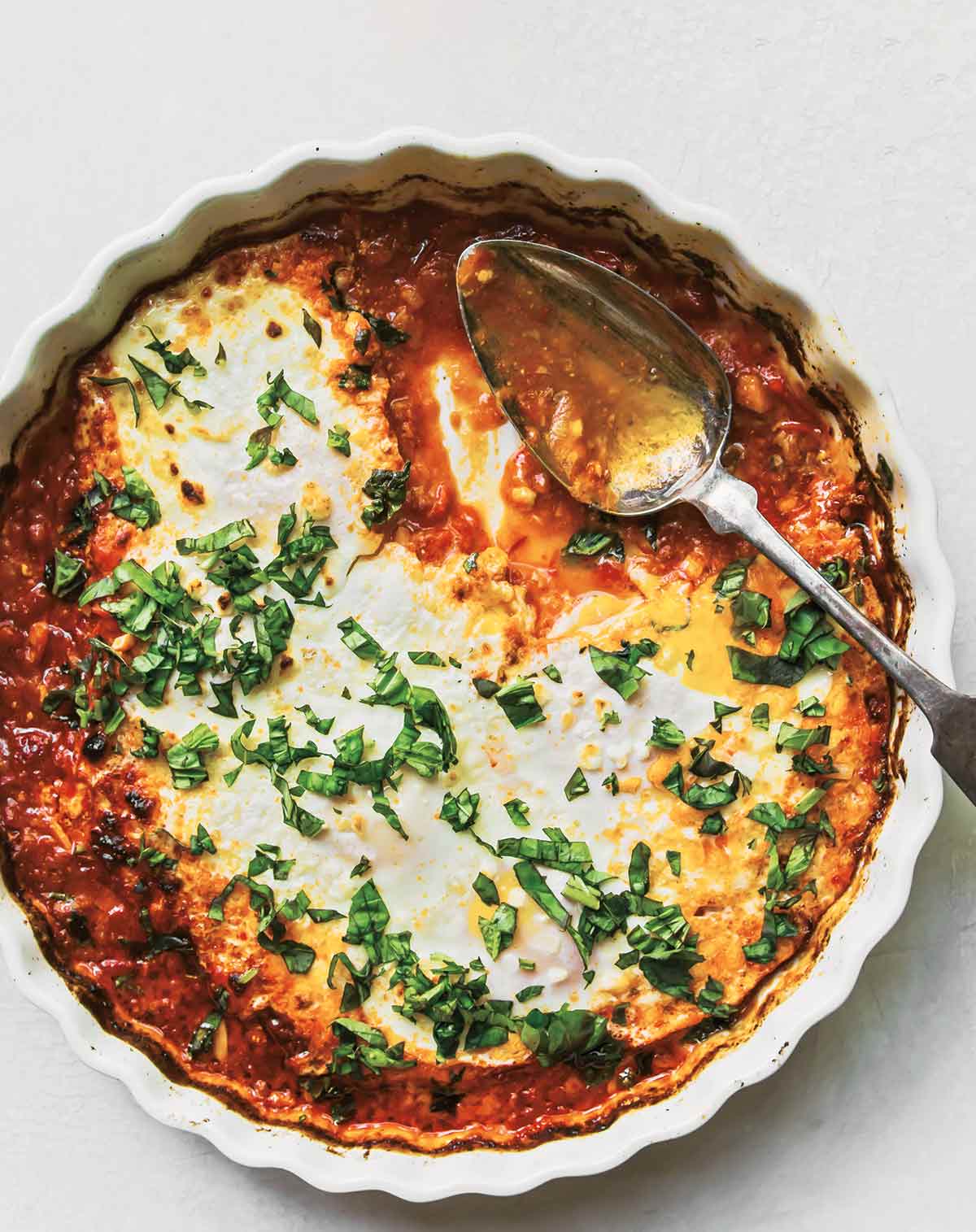 A white ribbed-edge tart pan filled with harissa baked eggs with goat cheese, with a spoon and topped with herbs.