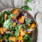 A wooden bowl filled with kale, butternut squash, and farro salad with a wooden spoon and kale and farro scattered on the tabletop.