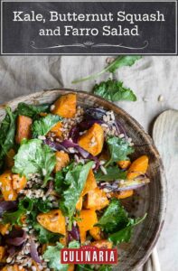 A wooden bowl filled with kale, butternut squash, and farro salad with a wooden spoon and kale and farro scattered on the tabletop.