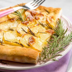 A purple napkin with a white plate on top, with a slice with potato rosemary focaccia garnished with rosemary and with a fork on top.