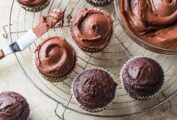 Vegan chocolate cupcakes, half frosted with vegan chocolate frosting, on a wire rack beside a bowl of frosting.