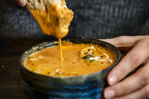 Butternut squash soup in a blue pottery bowl with a slice of rustic bread being dipped into it.