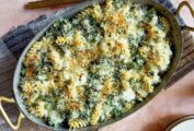 Baked cheesy pasta with broccoli in a large metal casserole dish, with a serving spoon and napkin.