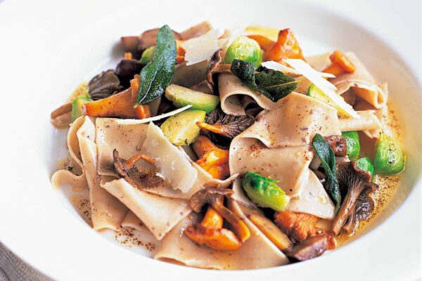 Chestnut pasta with wild mushrooms and Brussels sprouts on a white plate garnished with sage leaves.