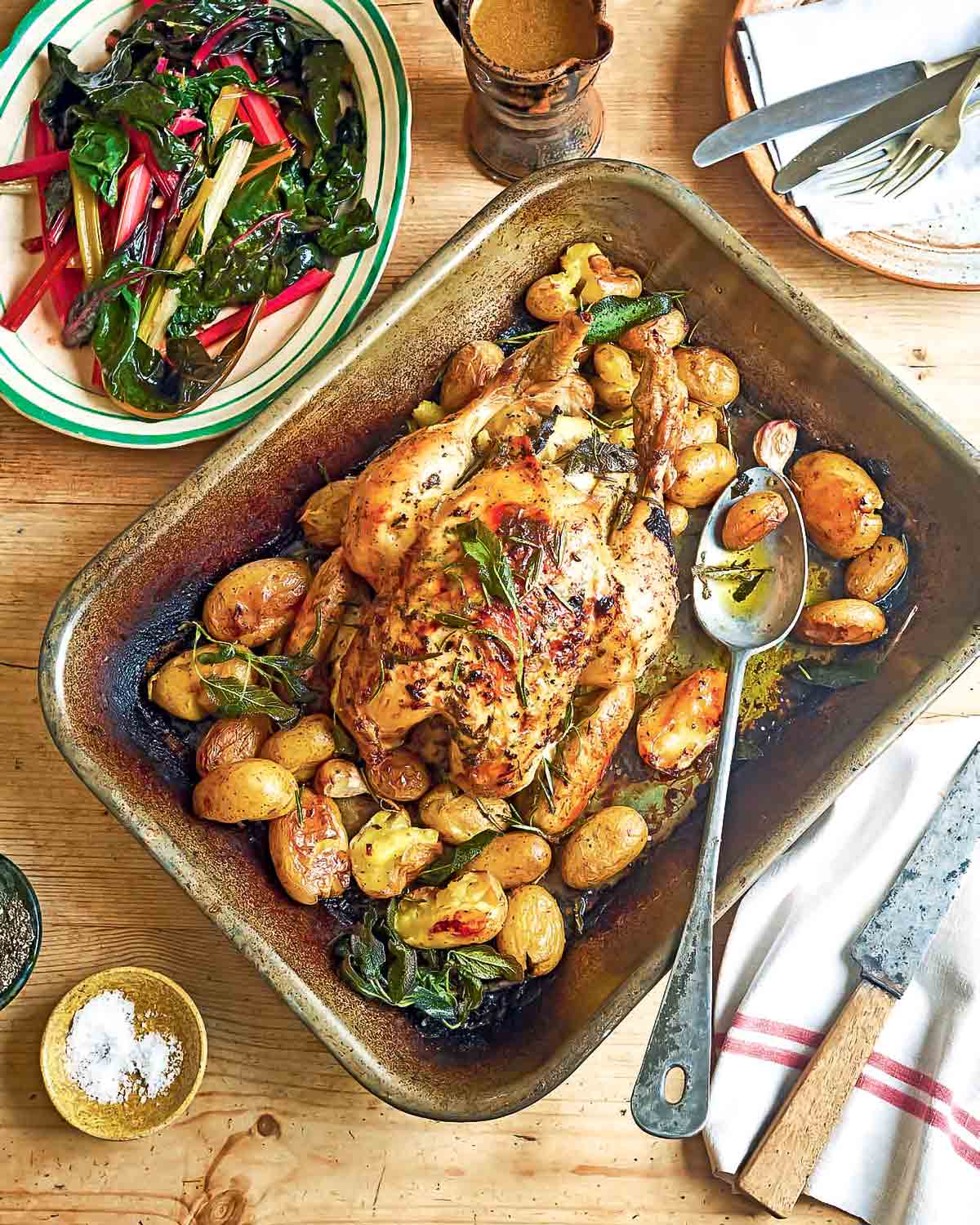 Lemon and herb roast chicken with fingerling potatoes and cloves of garlic, in a roasting pan, beside a plate of Swiss chard salad.
