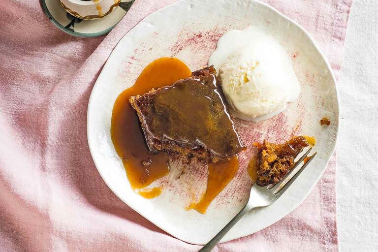 Sticky toffee pudding with vanilla ice cream on a white plate with a fork, beside a small bowl of extra toffee sauce.