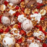 Banana split nachos on a large baking sheet, garnished with almonds, cherries, and chocolate sauce.