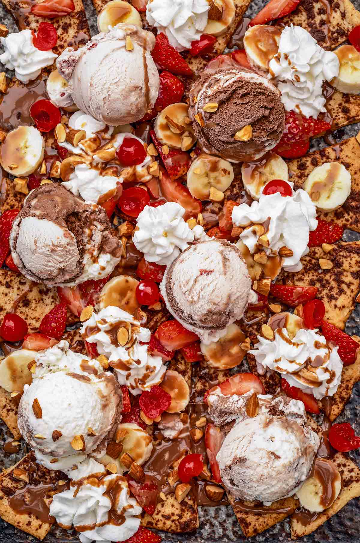 Banana split nachos on a large baking sheet, garnished with almonds, cherries, and chocolate sauce.