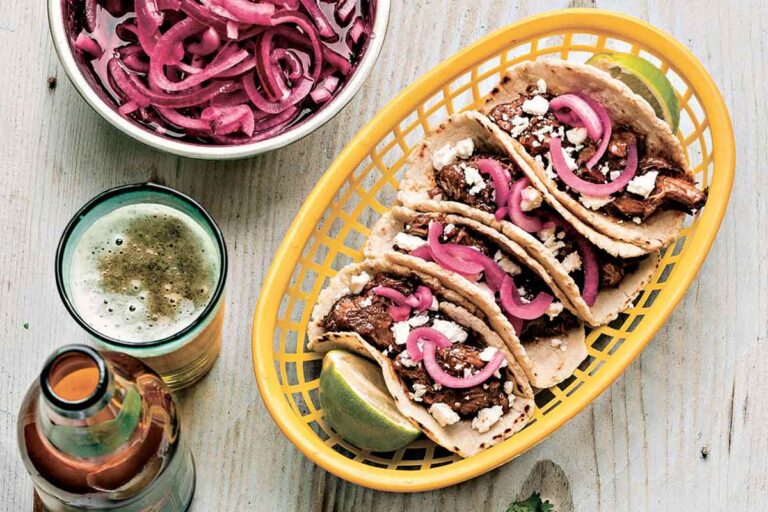 Four beef chili tacos and two lime halves in a basket, with bowls of pickled onions, slaw, and a beer on the side.