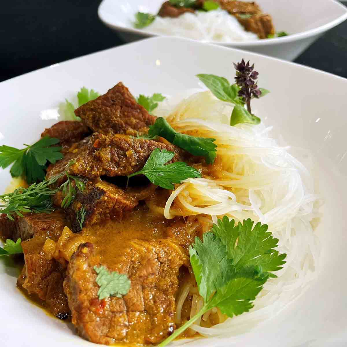 Burmeses beef curry in a white bowl with vermicelli noodles and cilantro and basil garnish.