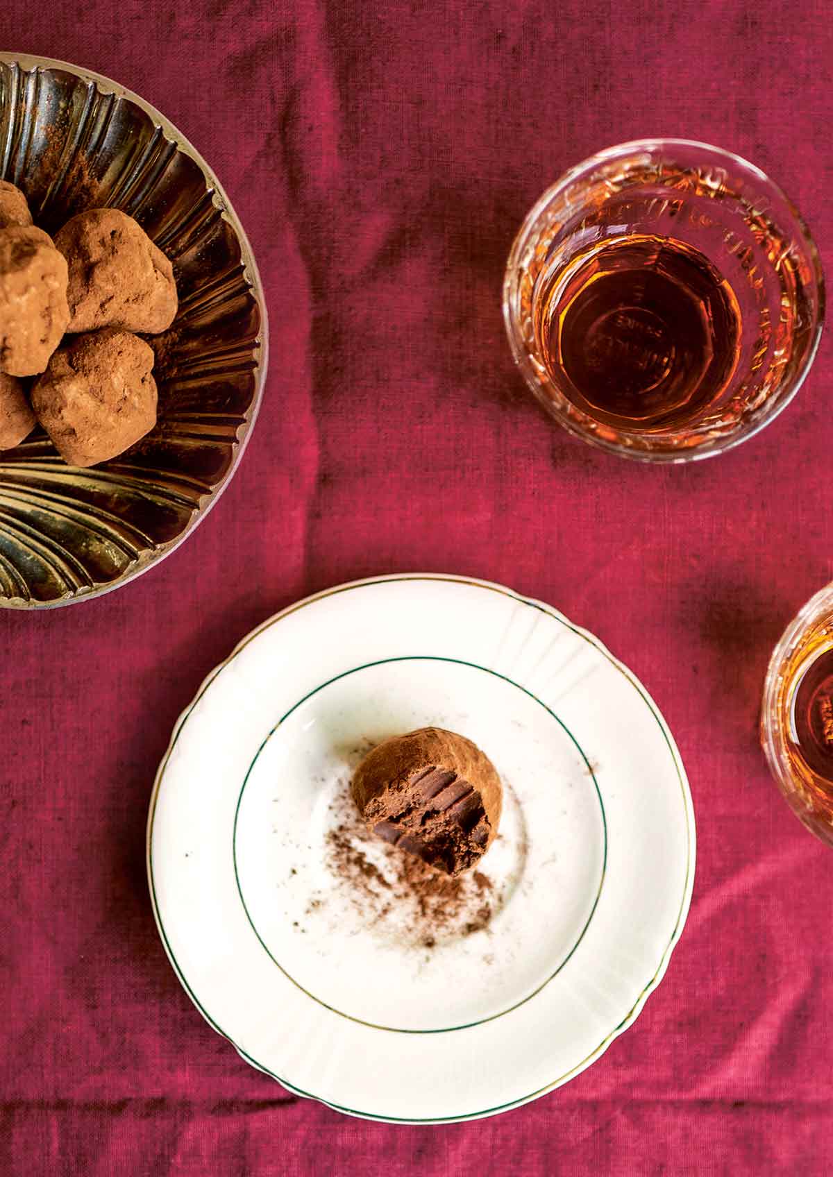A chocolate salted caramel truffle with a bite taken out of it, on a white saucer, beside a bowl of truffles and two glasses.