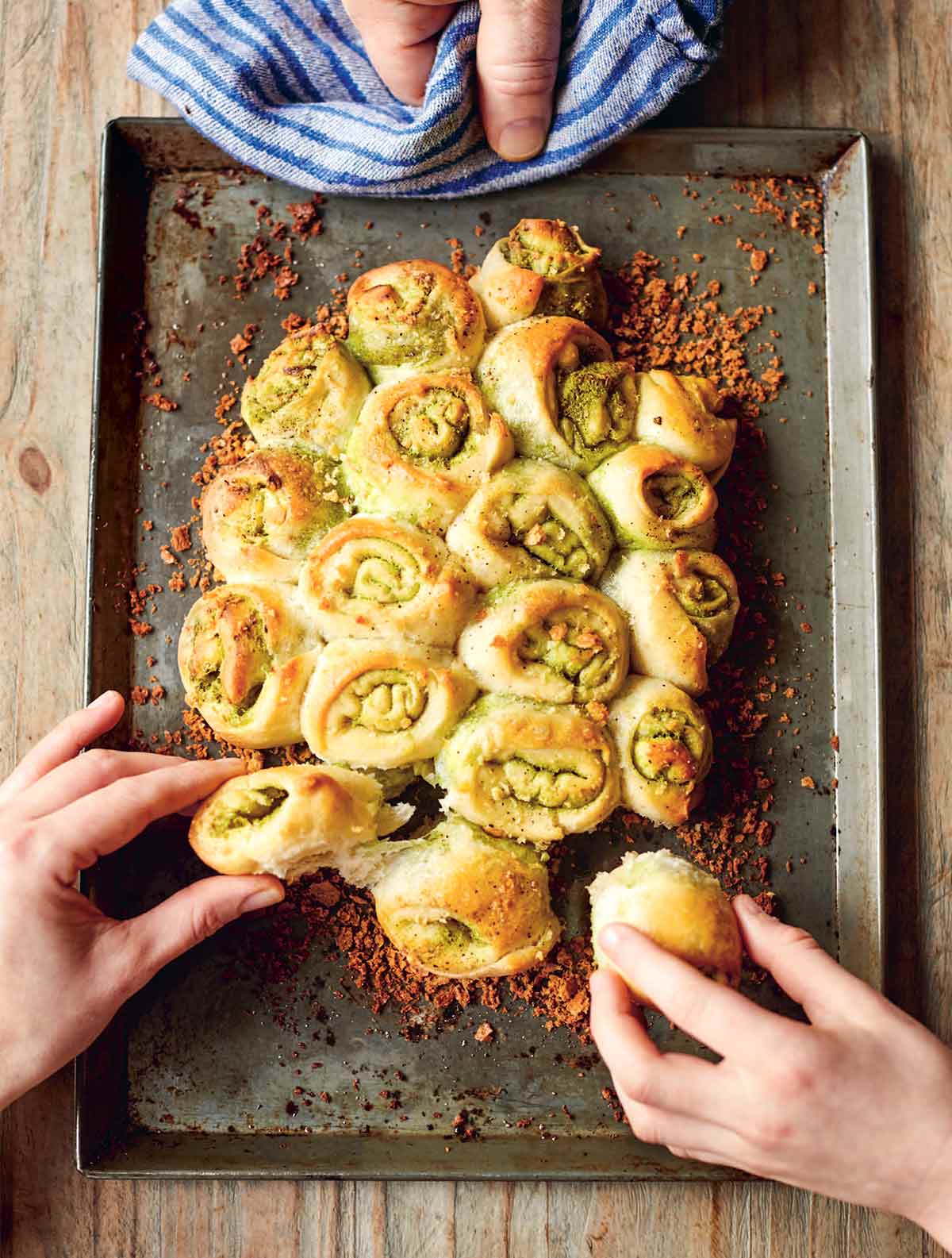 Jamie Oliver garlic rolls on a metal sheet pan, garnished with crispy bread crumbs, and being pulled apart by two hands.