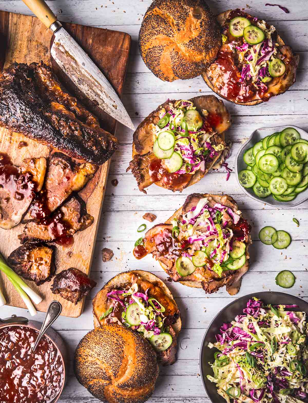 Four Korean-inspired brisket sandwiches with sesame coleslaw on a table, with bowls of coleslaw, sliced cucumbers, gochujang, and a cutting board with sliced brisket.