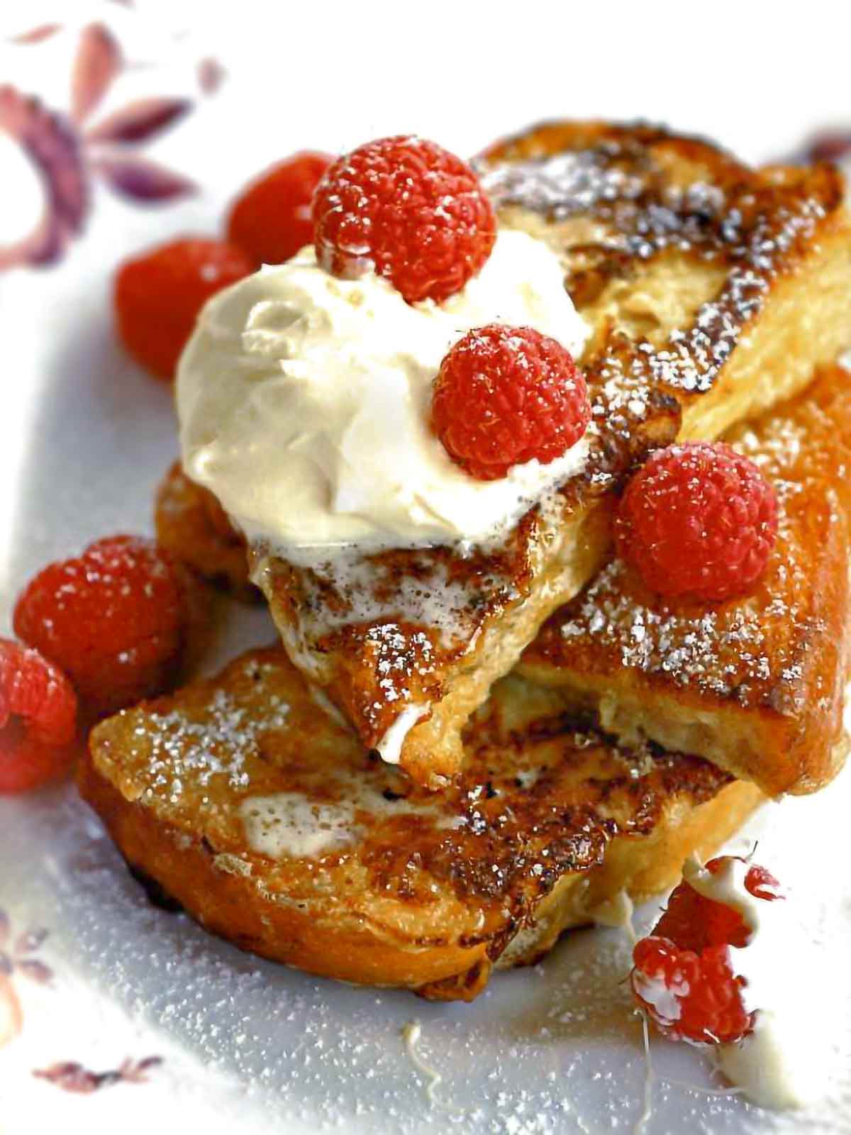 Pain perdu-French toast with raspberries piled on a plate dusted with icing sugar, a dollop of whipping cream, and raspberries.