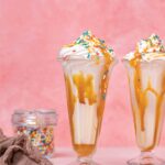 Salted caramel funfetti milkshakes in tall parfait glasses with sprinkles, beside a jar of sprinkles and a linen napkin.