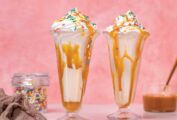 Salted caramel funfetti milkshakes in tall parfait glasses with sprinkles, beside a jar of sprinkles, a linen napkin, and a jar of caramel sauce.