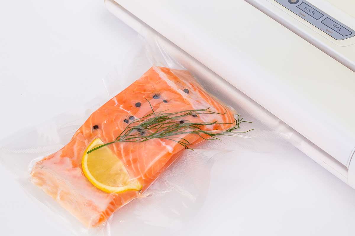 A salmon filet in a plastic bag with peppercorns, a lemon wedge and dill sprig, being vacuum sealed.