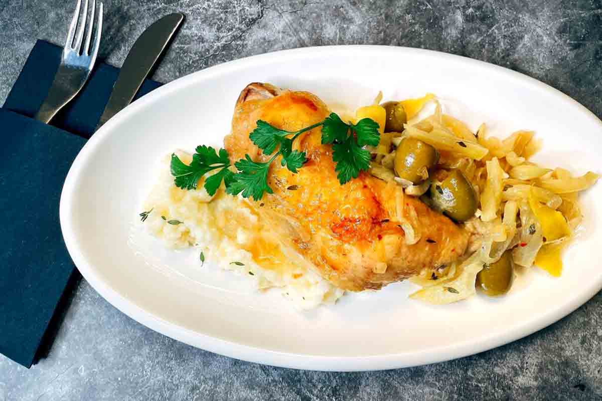 A plate with a leg and though of Mediterranean chicken with fennel and green olives sitting on a bed of mashed potatoes.