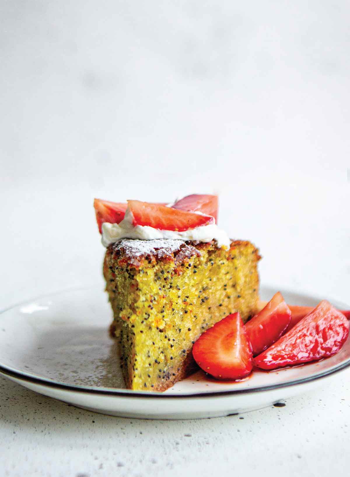 A slice of orange, olive oil, and poppyseed cake with sliced strawberries on the side on a white plate